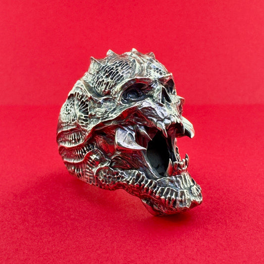 A highly detailed, open-mouthed dragon skull ring made of textured silver-toned metal, set against a solid red background.