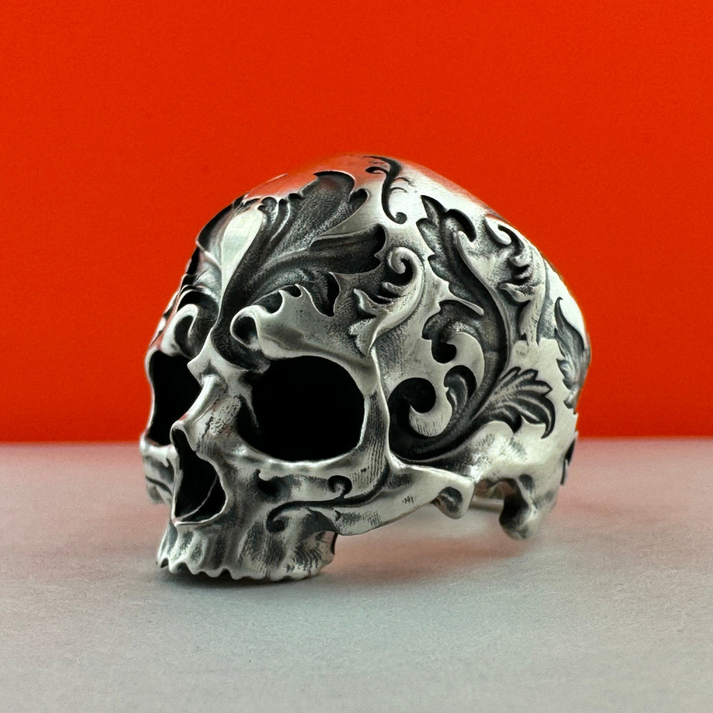 A silver skull ring with intricate baroque-style filigree and scrollwork, photographed on a light gray surface against a vibrant red-orange background.